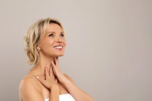 Double Chin Treatment. Beautiful Middle Aged Woman Touching Soft Smooth Skin On Neck, Attractive Mature Lady Standing Wrapped In Towel OverGrey Background, Enjoying Her Beauty, Copy Space